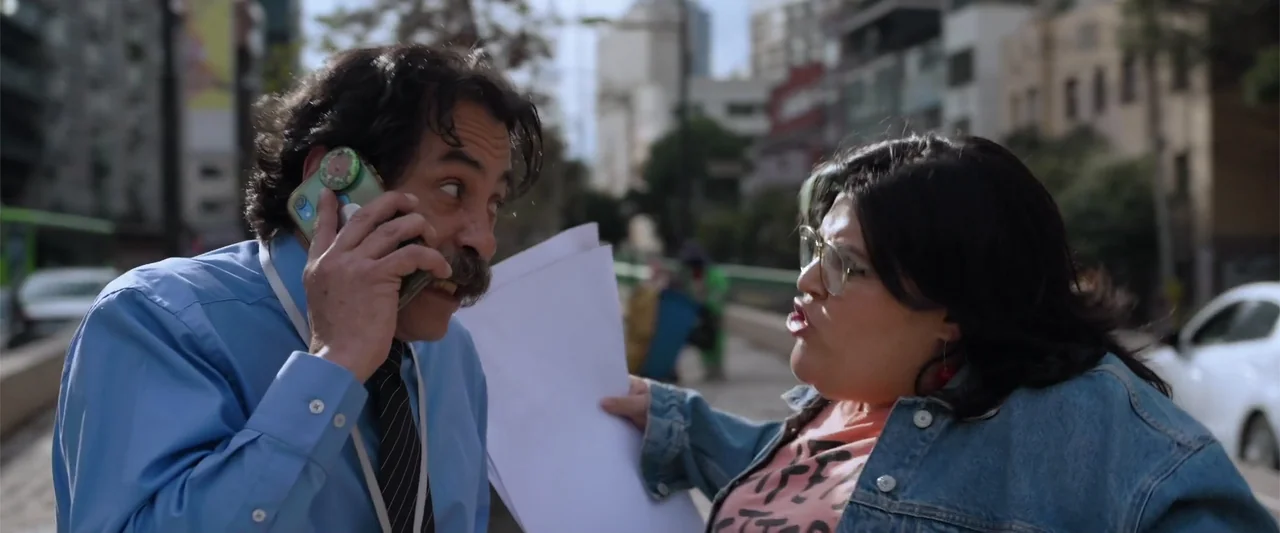 Winner Takes the Cake (2025) movie still: medium — An urban outdoor sidewalk with blurred city buildings visible in the background. A man on a phone…