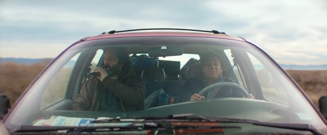 The Long Dumb Road (2018) movie still: medium — The interior of a moving car showing two men driving through an open landscape. The driver holds the…