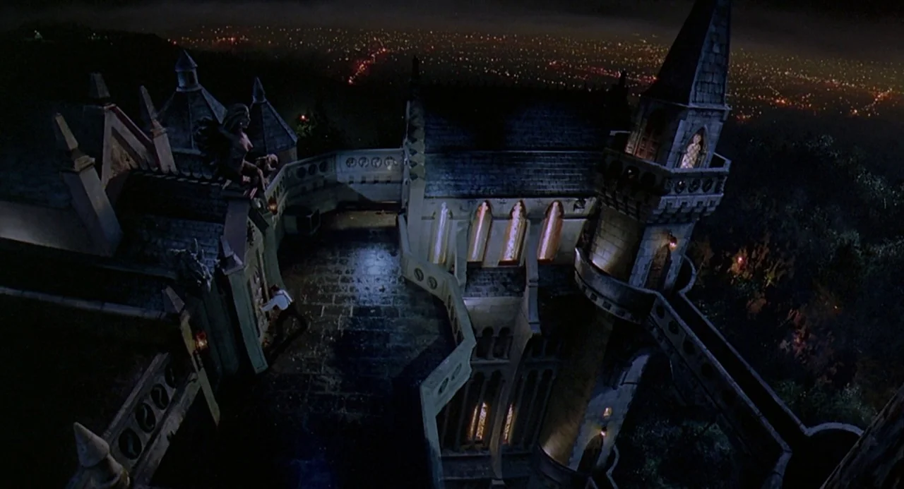 Death Becomes Her (1992) movie still: establishing — A high-angle view looks down onto the intricate stone roof of a large mansion. In the distance,…