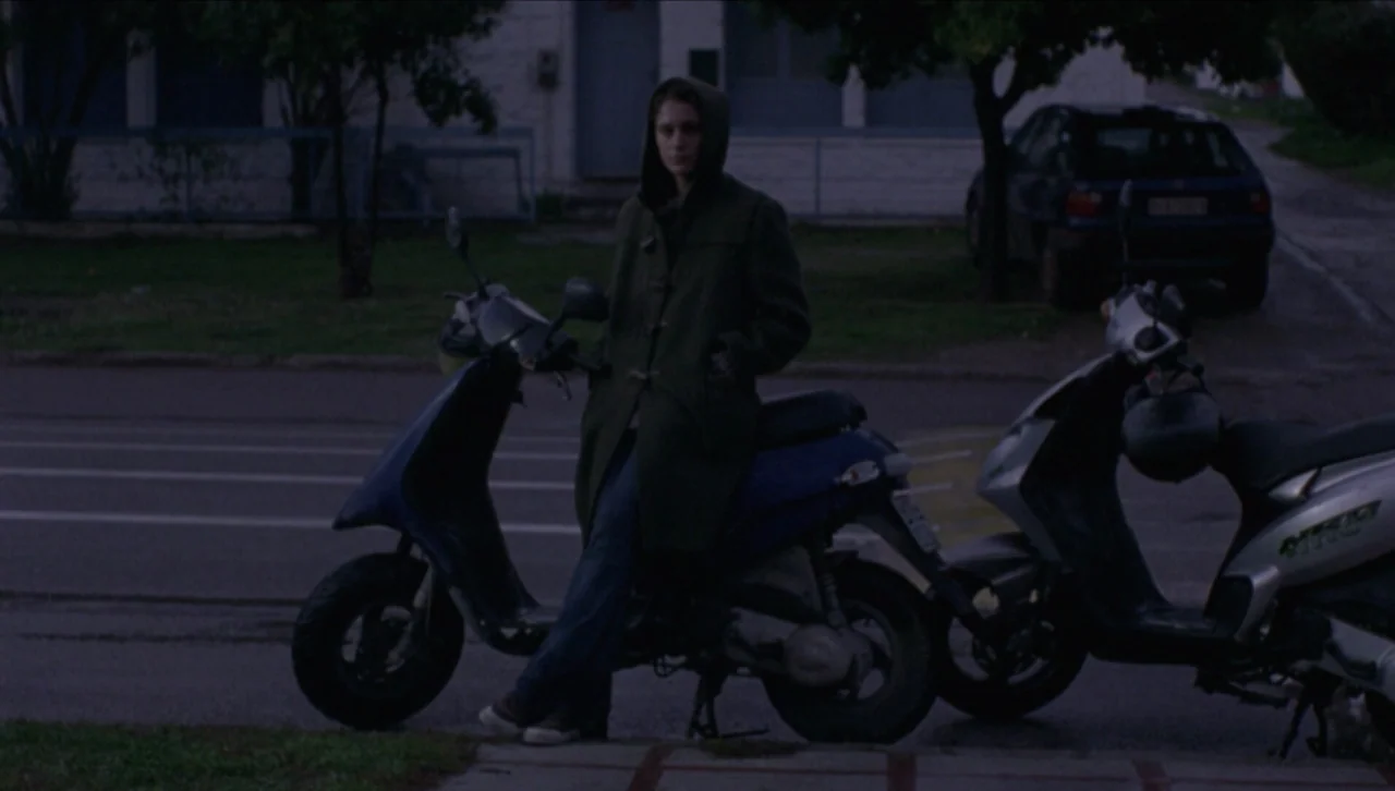 Attenberg (2010) movie still: fullbody — A person stands on a residential sidewalk at night beside two parked motor scooters. The background shows a…