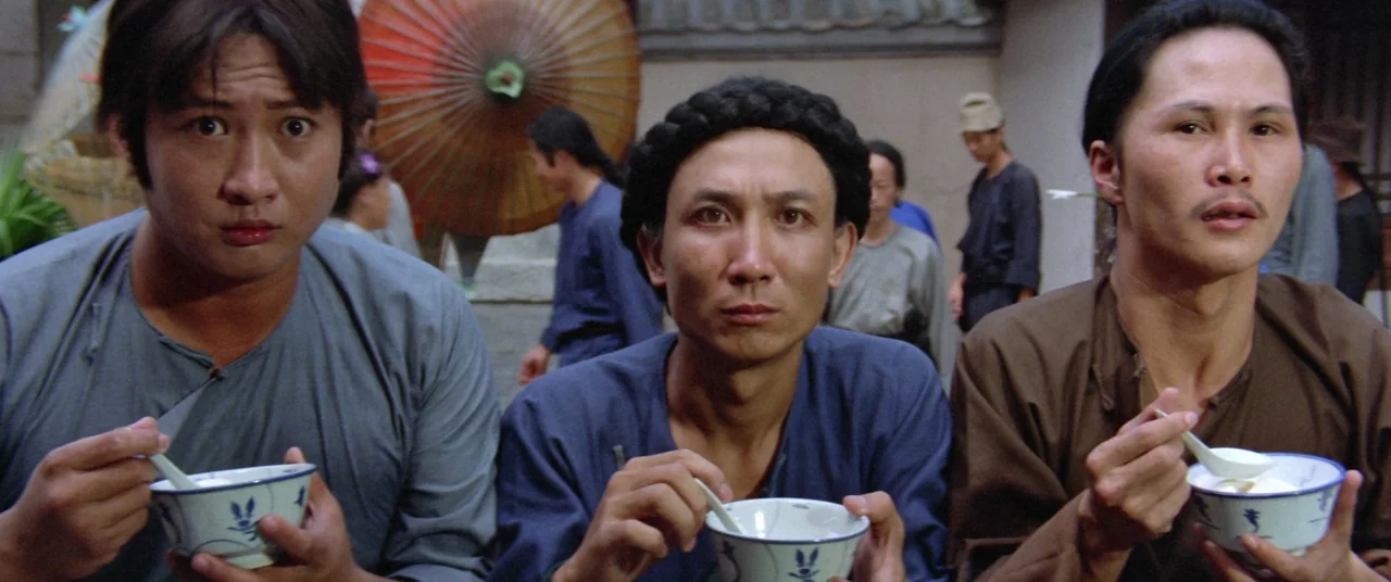 Encounter of the Spooky Kind (1980) movie still: medium — Three men stand side by side in a busy outdoor market environment. Each man holds a small…