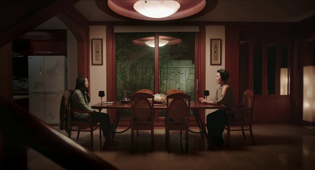 Microhabitat (2017) movie still: wide — A dimly lit dining room features wood-paneled walls and a central rectangular table. Two women sit facing…