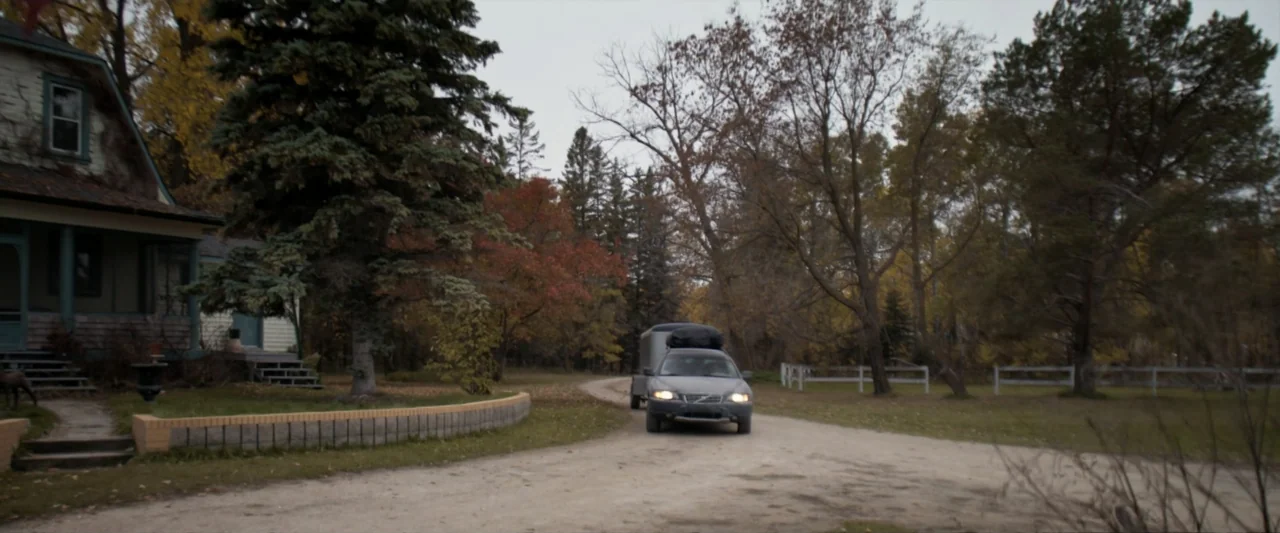 Clown in a Cornfield (2025) movie still: wide — A gravel driveway leads toward a house framed by autumn trees under gray skies. A silver station…