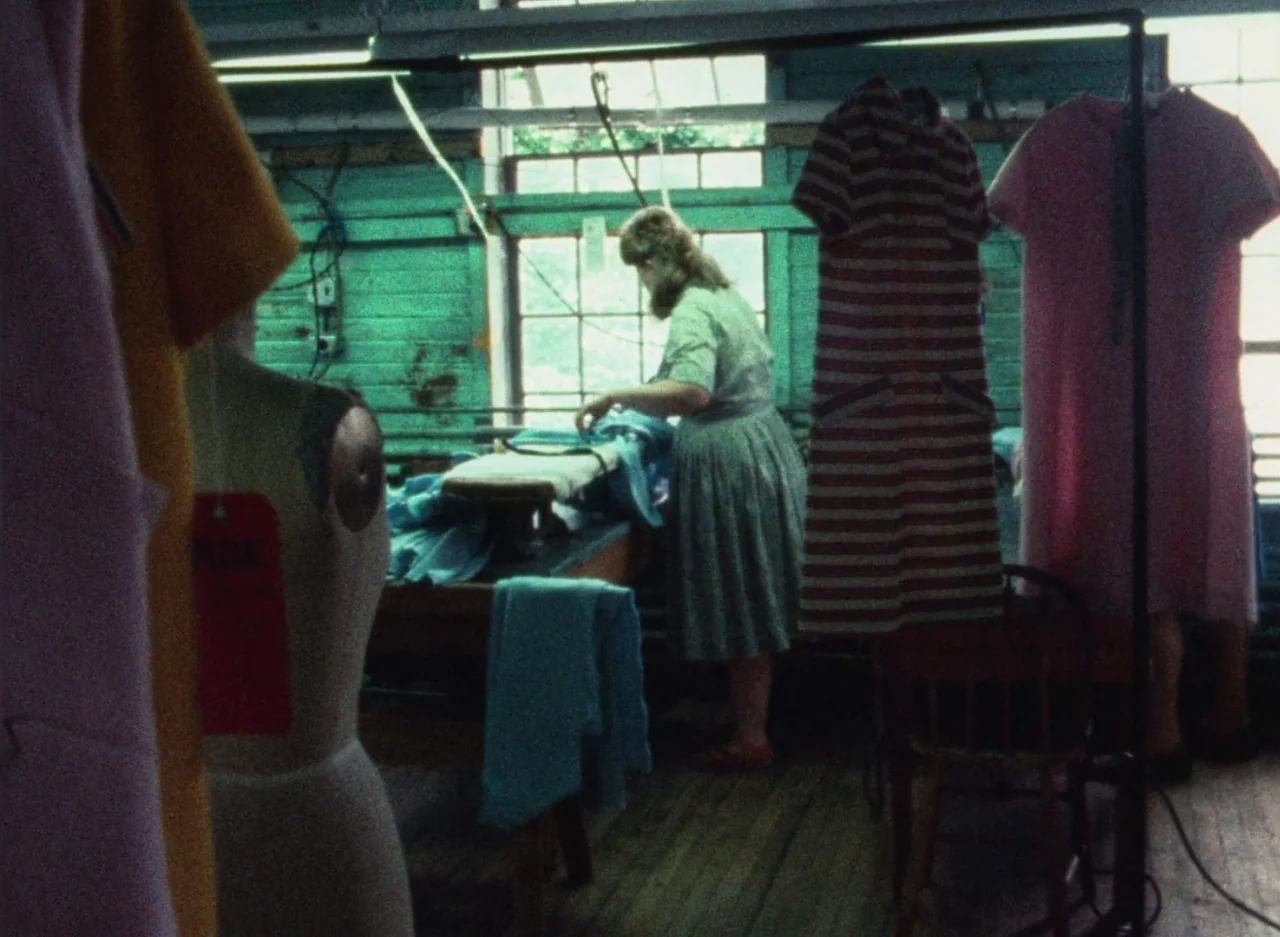 Wanda (1970) movie still: medium — A workspace with teal-painted wooden walls and hanging garments on racks. A woman works at an ironing board near…