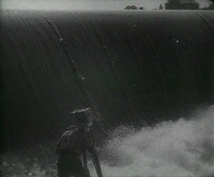 Man with a Movie Camera (1929) movie still: wide — A man stands before a large curved concrete dam with water pouring over it. The filmmaker…