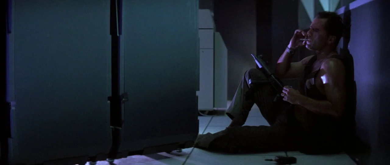 Die Hard (1988) movie still: medium — A dark, cramped industrial crawlspace with metal panels and low lighting. A man sits on the floor resting…