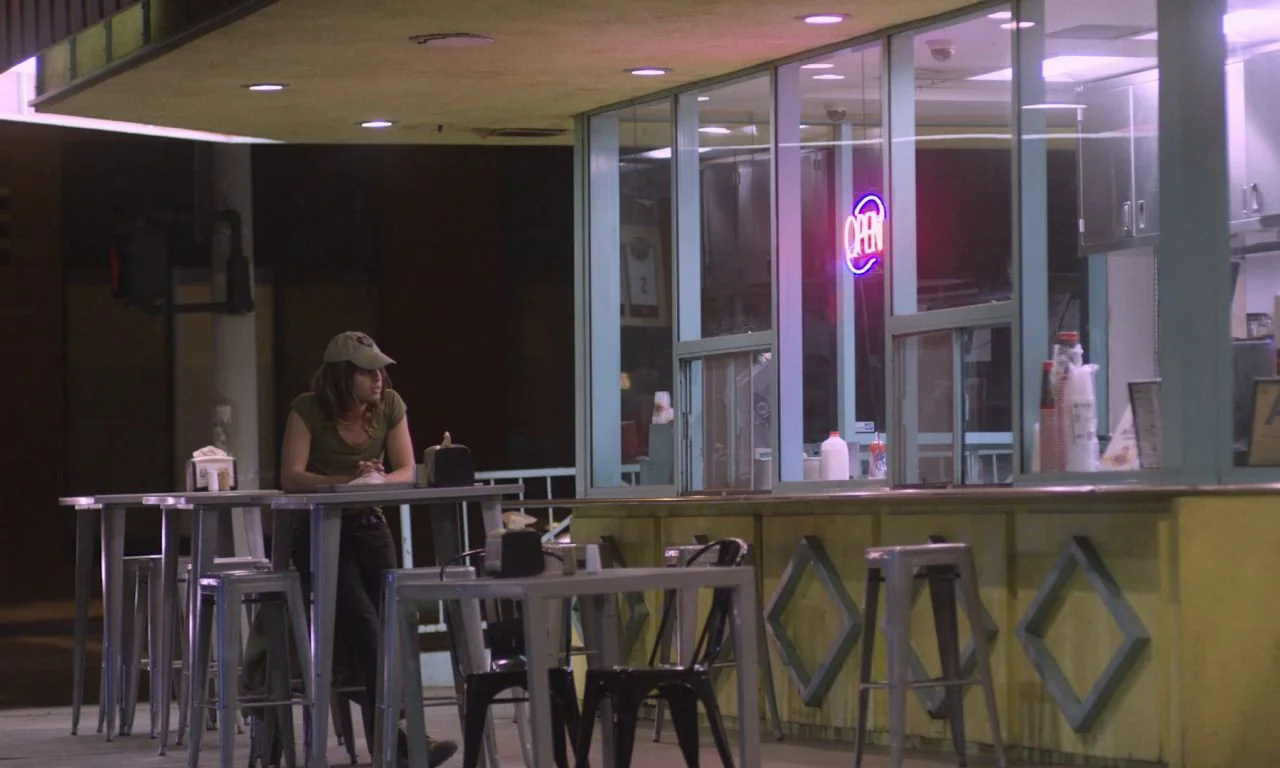 Ham on Rye (2019) movie still: wide — A nighttime exterior of a fast-food outlet featuring yellow walls and large glass windows. A young man sits…