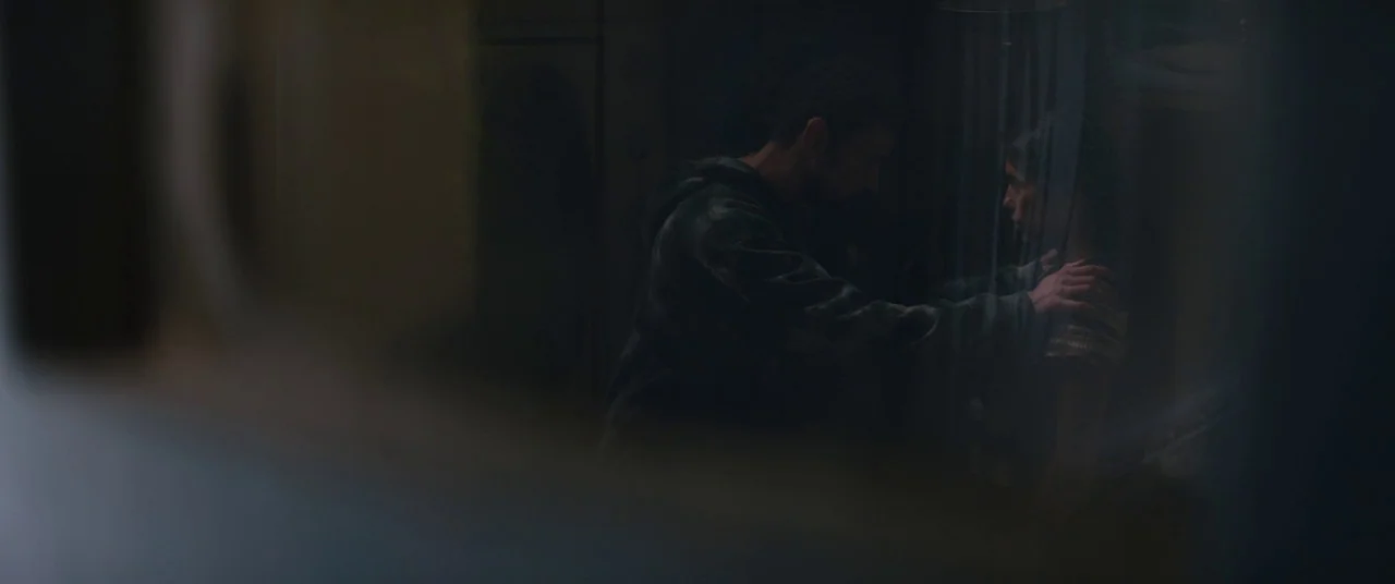 The Rhythm Section (2020) movie still: medium — A dimly lit, shadowed room with indistinct walls and dark surroundings. A man faces a woman and…