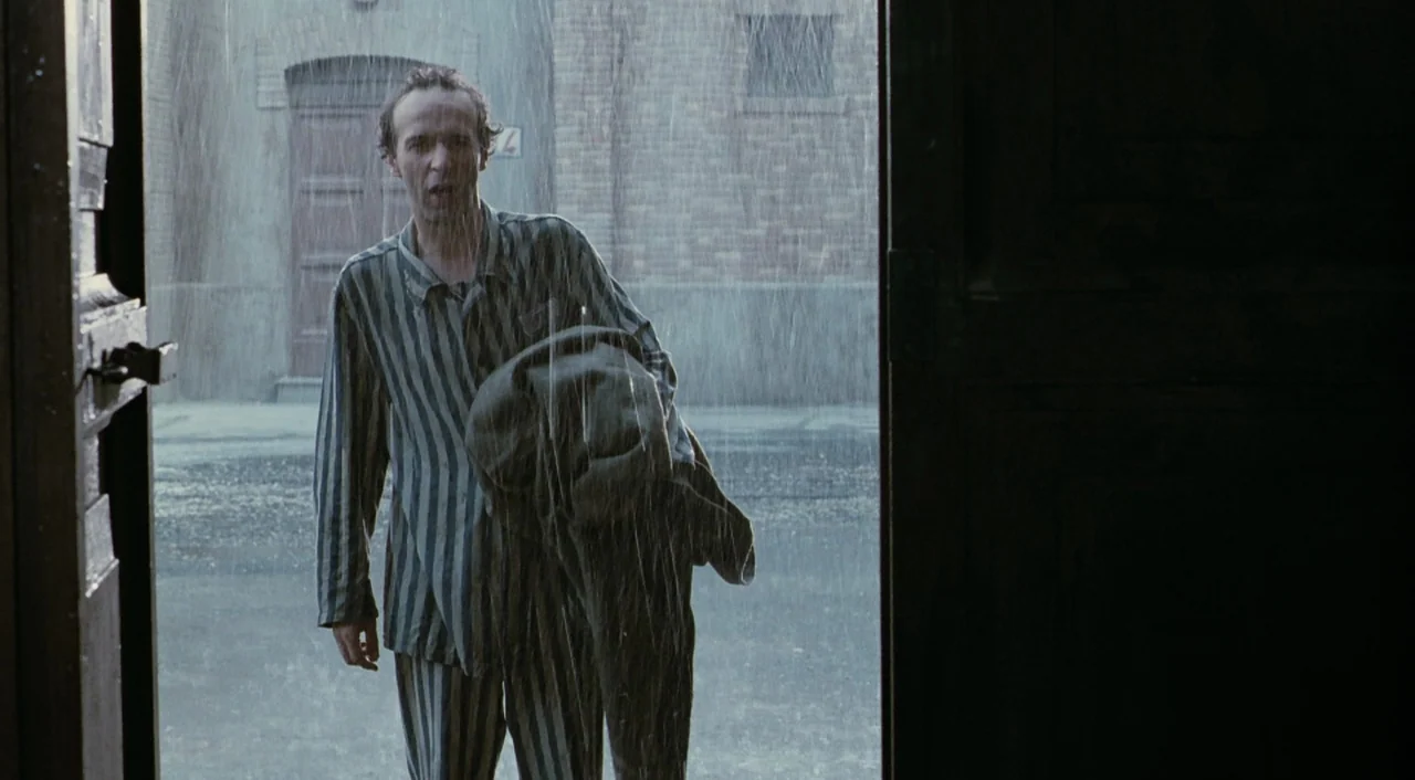 Life is Beautiful (1997) movie still: medium — An outdoor prison compound with brick buildings is obscured by a heavy downpour. A man wearing…