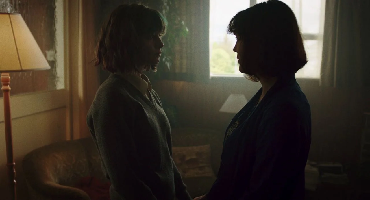 Dating Amber movie still: medium — A dimly lit room with a window providing backlight through the frame. Two women stand facing each other in the…