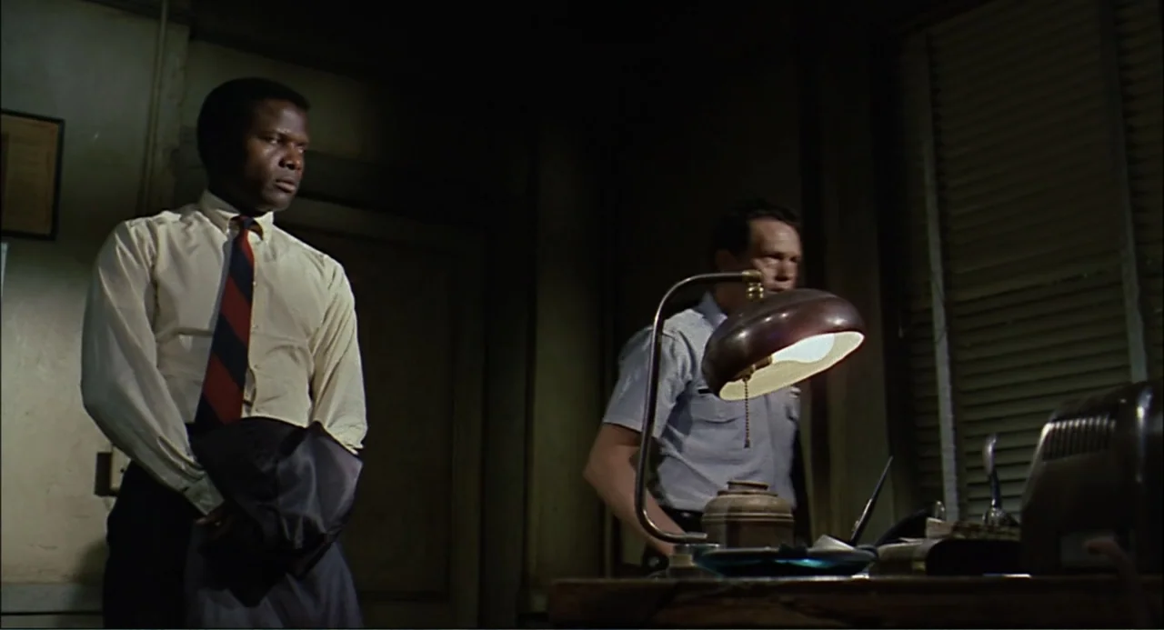 In the Heat of the Night (1967) movie still: medium — A dimly lit room features a wooden desk with a metal lamp. One man stands while another man…