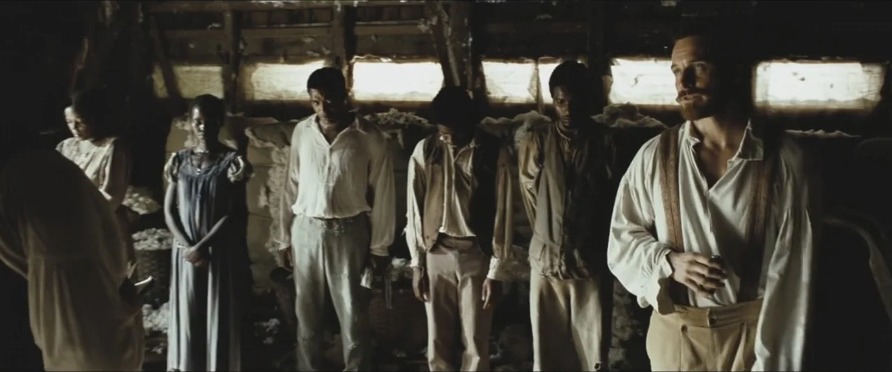 12 Years a Slave (2013) movie still: fullbody — A dimly lit wooden building contains raw cotton piles and several standing figures. A group of…
