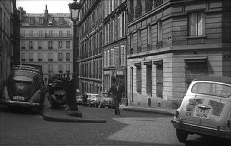 Le Signe du Lion (1962) movie still: wide — A narrow European city street flanked by tall, multi-story limestone apartment buildings with shutters.…