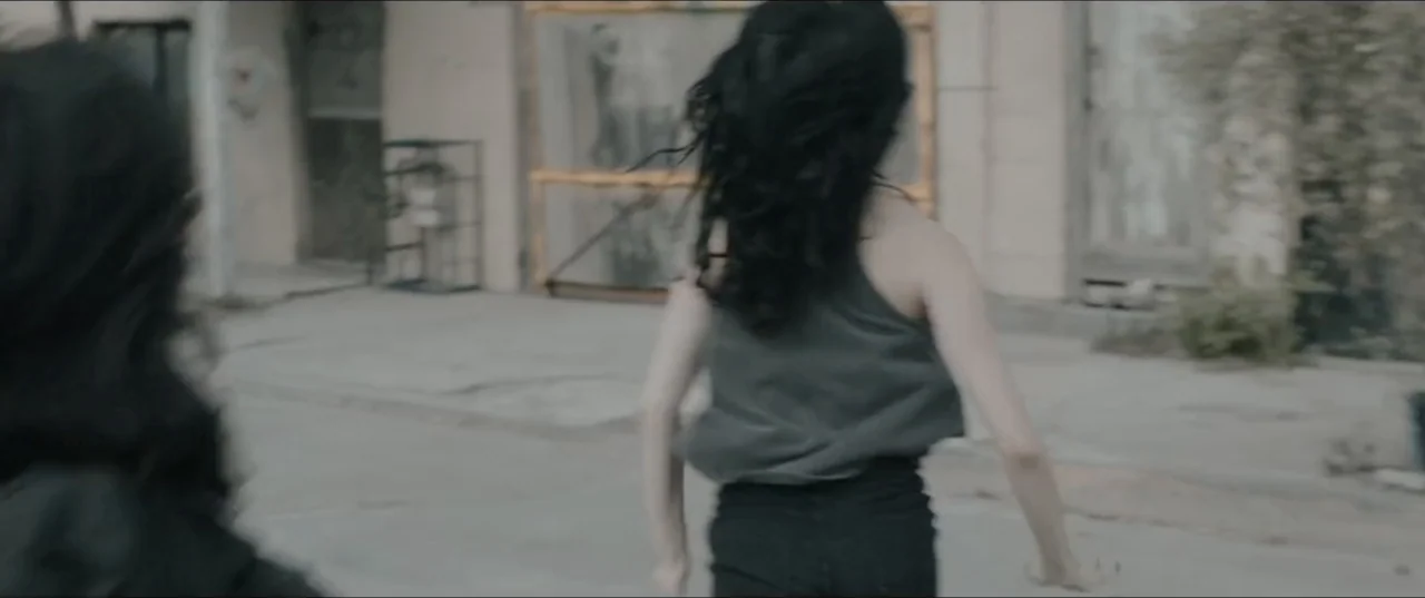 Death to Summer (2021) movie still: medium — An urban outdoor street scene with weathered buildings and light-colored concrete ground. Two young…