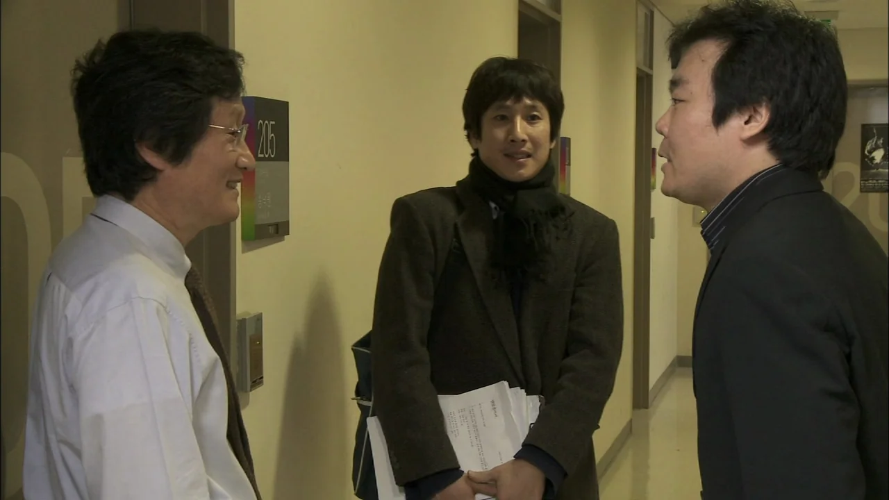 Oki’s Movie (2010) movie still: medium — An office hallway with beige walls and numbered doors displays a small room sign. Three men stand in a…