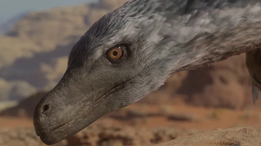 Prehistoric Planet (2022) movie still: closeup — A feathered dinosaur head occupies the foreground of a vast, arid sandy desert landscape. The…