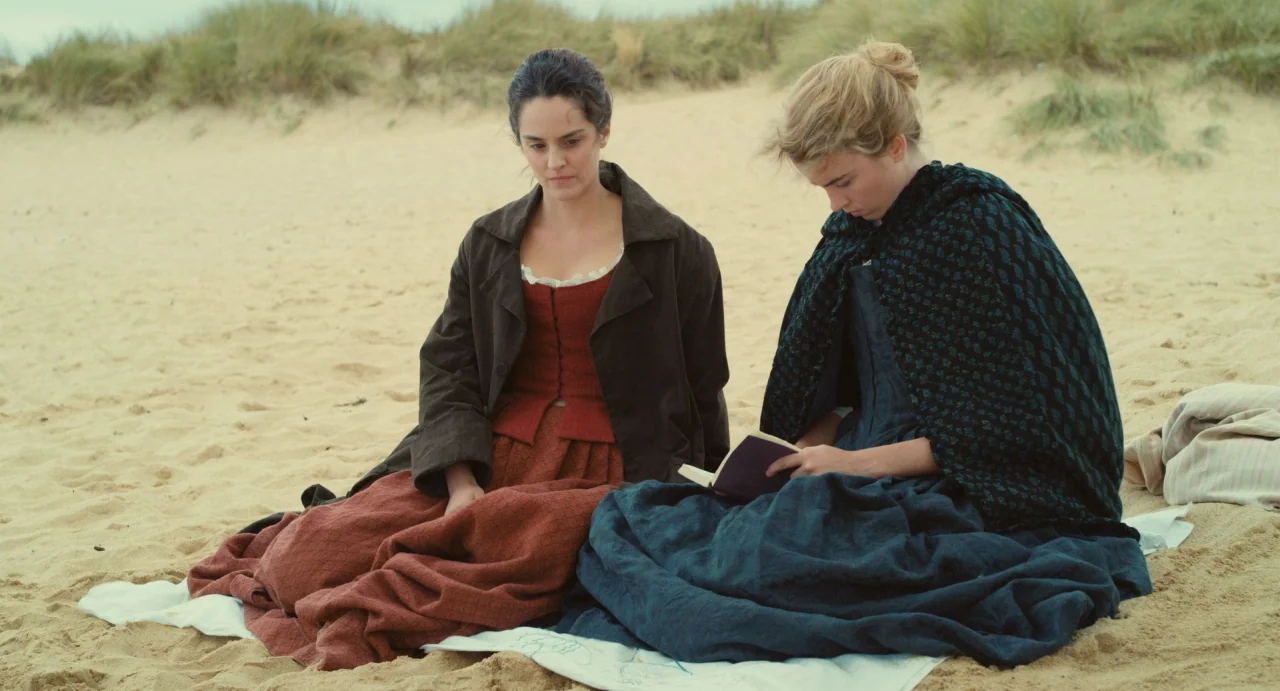 Portrait of a Lady on Fire movie still: fullbody — Two women in 18th-century attire sit on a white cloth on a sandy beach. One woman reads a book…