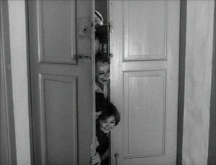 Even Dwarfs Started Small (1970) movie still: medium — A vertical stack of people peer through a narrow gap between two wooden doors. The subjects…