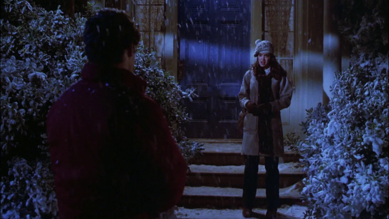 Gremlins (1984) movie still: medium — A dark house entrance at night features a blue door framed by snow-covered bushes. A woman in winter gear…