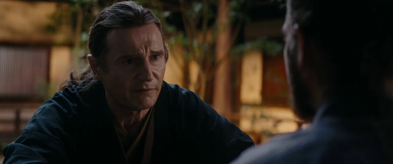 Silence (2016) movie still: medium — A dimly lit room with blurred architectural structures and vegetation visible in the background. A man with…
