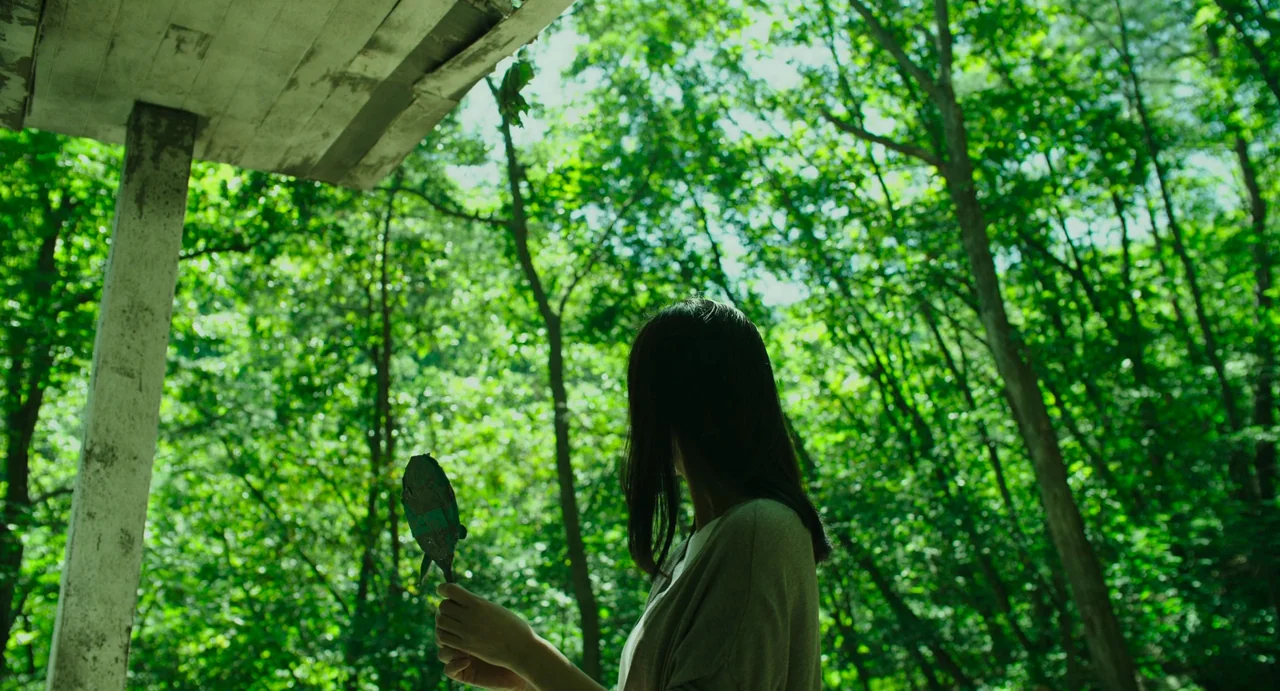 The Mimic (2017) movie still: medium — A woman stands under a wooden overhang looking toward a dense green forest. She holds a small, weathered…
