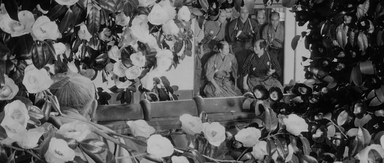 Sanjuro (1962) movie still: wide — A group of samurai men wait in a wooden building, viewed through thick camellia bushes. The foreground is heavily…