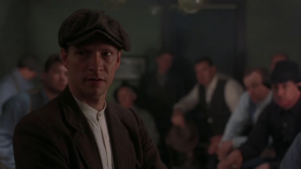 Matewan (1987) movie still: medium — A dimly lit room filled with men sitting in a semi-circle during the 1920s. The main subject in a newsboy cap…