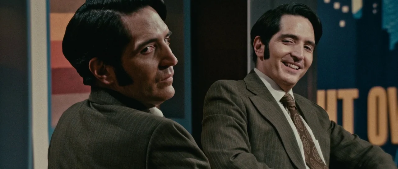 Late Night With The Devil (2023) movie still: medium — A televised set features retro orange and blue graphic panels in the background. A man in a…