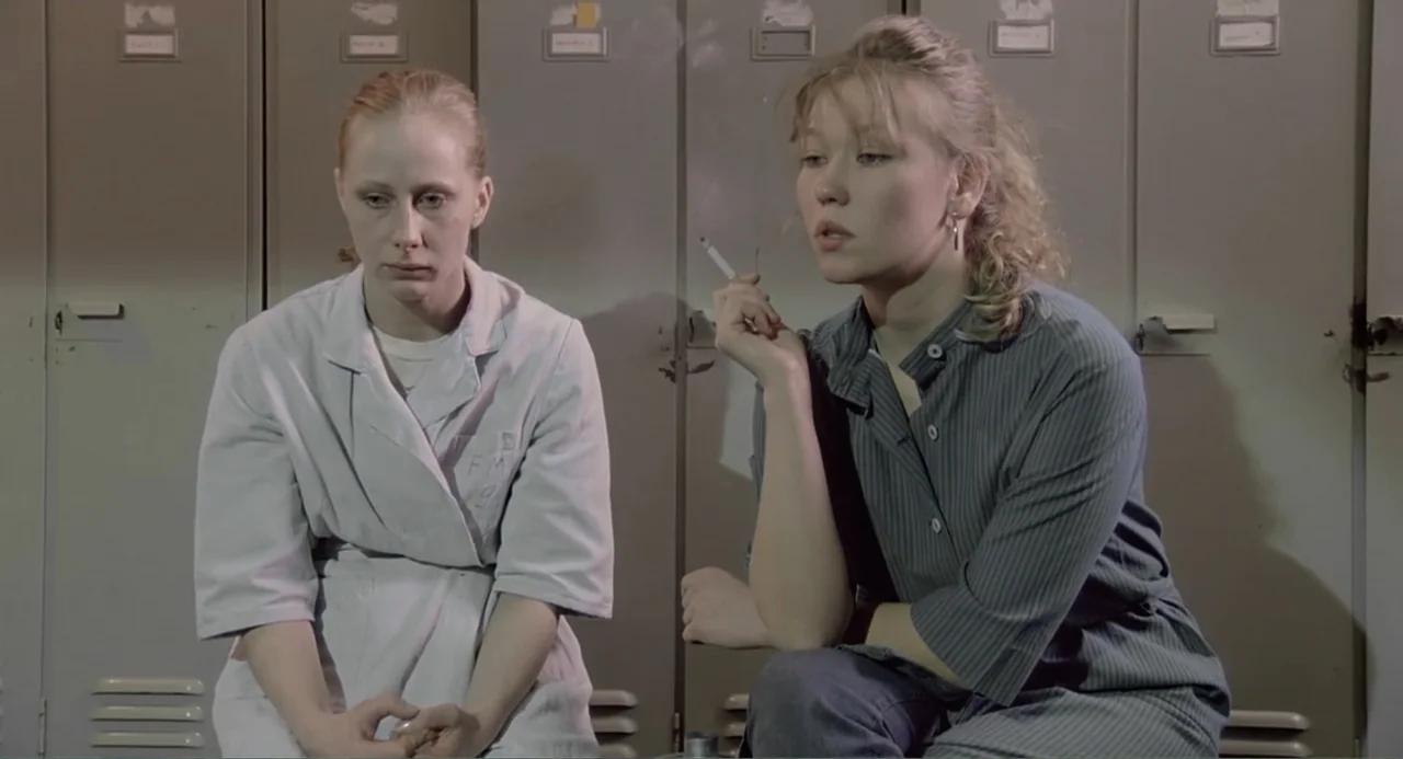 The Match Factory Girl (1990) movie still: medium — A row of gray metal lockers lines the background wall in a utilitarian space. Two women sit in…