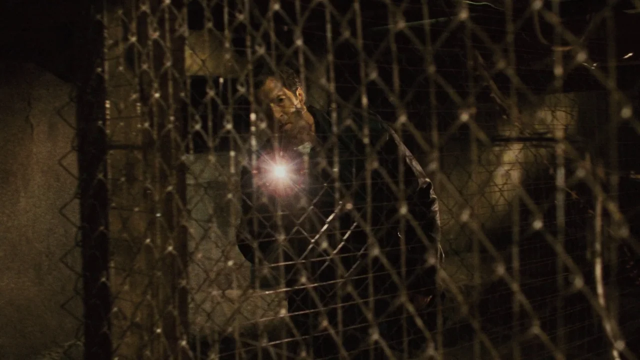 Saw V (2008) movie still: medium — A dimly lit industrial basement featuring a prominent chain-link fence as a foreground element. A man behind the…