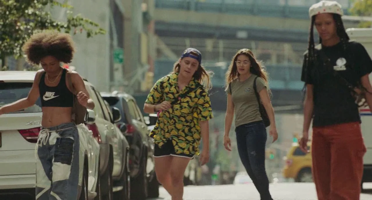 Skate Kitchen (2018) movie still: fullbody — Four teenagers walk across a sunlit city street lined with parked vehicles. The group moves together…