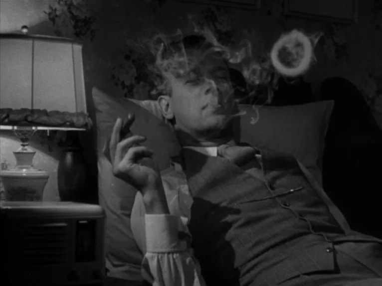 Shadow of a Doubt (1943) movie still: medium — A man reclines against pillows on a bed with floral patterned wallpaper in the background. He holds a…