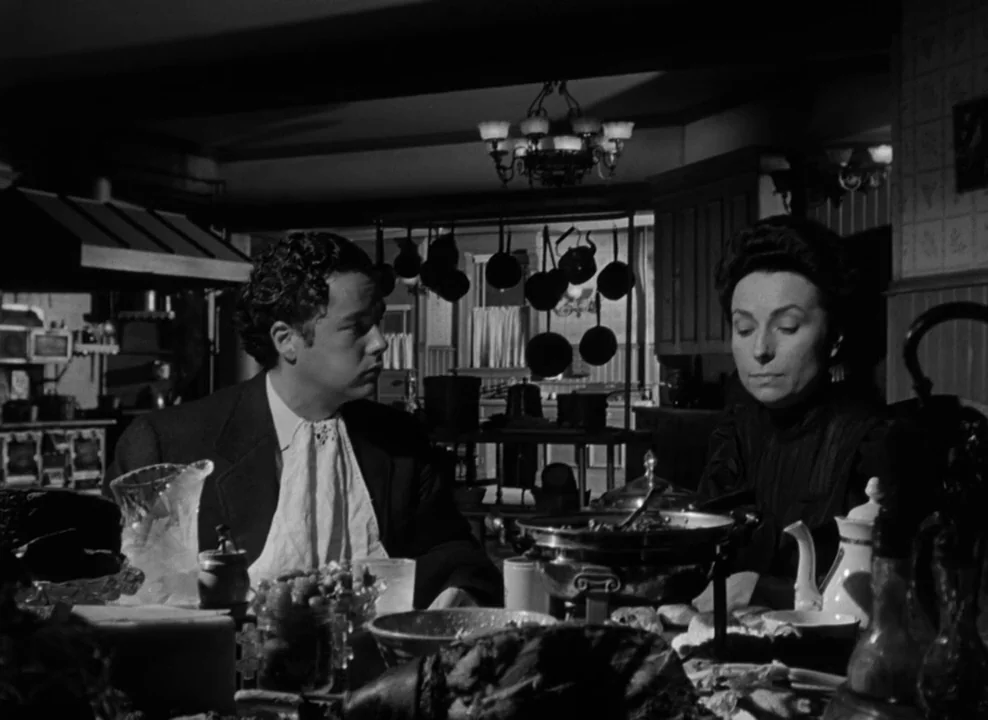 The Magnificent Ambersons (1942) movie still: medium — A dark, wood-paneled room features a table laden with serving dishes and dinnerware. A man…
