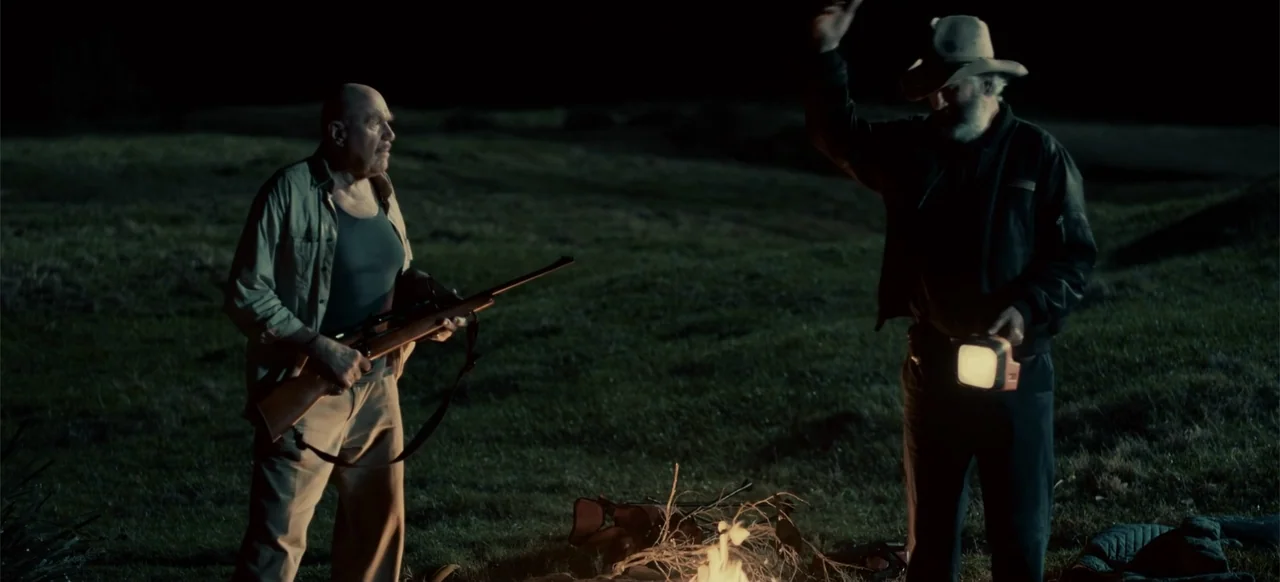 Boar (2018) movie still: medium — A dark, grassy field at night illuminated by a small, low-burning campfire. Two men stand near the fire, one…