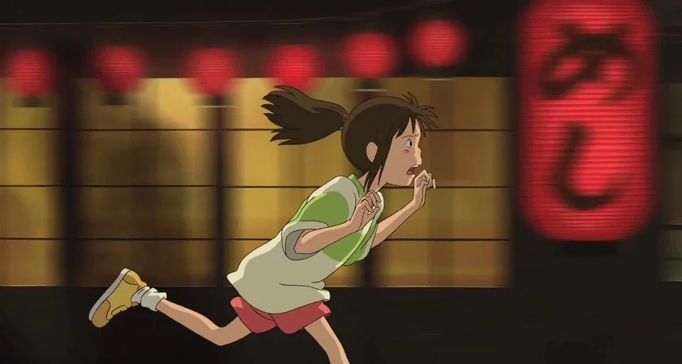 Spirited Away (2001) movie still: medium — A night street scene features a background of blurred horizontal red paper lanterns. A young girl with a…