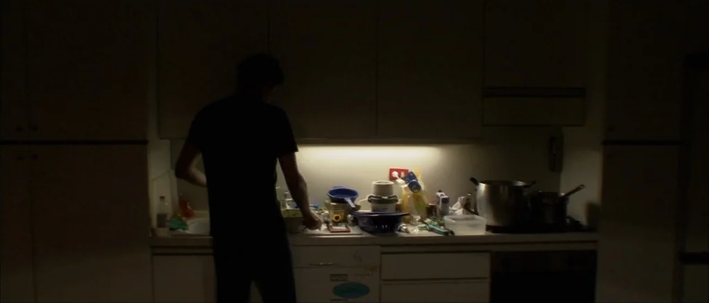 Genova (2008) movie still: medium — A dimly lit kitchen with cluttered countertops and overhead cupboards mostly obscured by shadow. A man stands…