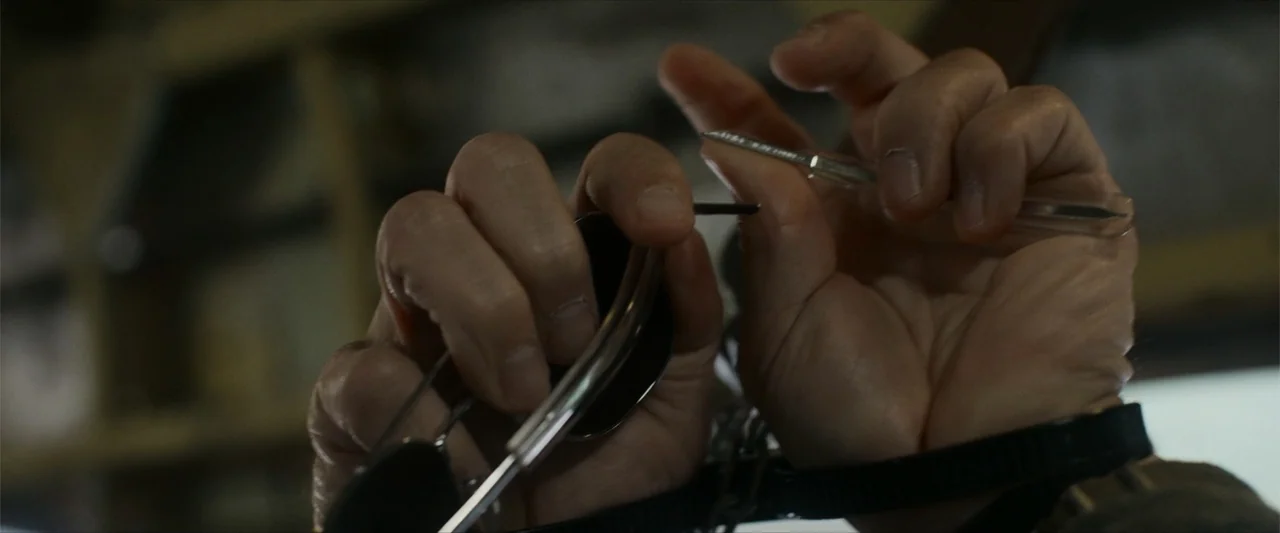 Flight Risk (2025) movie still: closeup — A close-up view shows a pair of hands working with metallic objects. The fingers manipulate a thin tool…