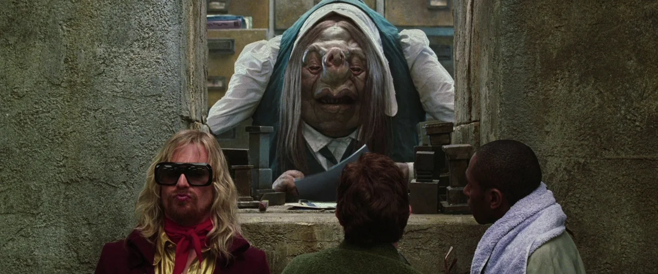 A Hitchhiker’s Guide To The Galaxy (2005) movie still: medium — A stone-walled office features a central desk where a creature manages paperwork.…
