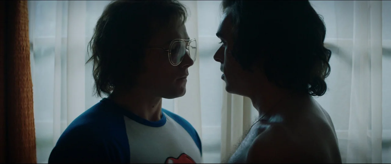 Rocketman (2019) movie still: medium — A residential room featuring a bright window covered by thin sheer white curtains. Two young men stand facing…