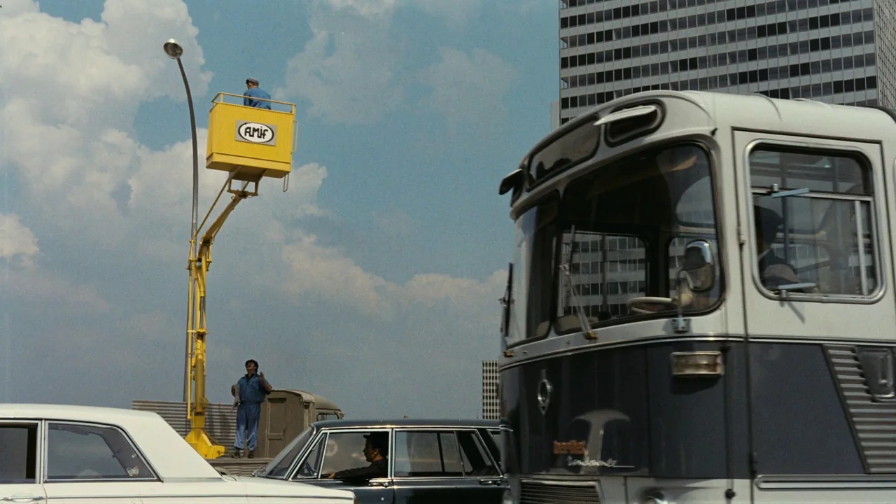 Playtime (1967) movie still: wide — A bright day on a multi-lane roadway with traffic and a large bus. A yellow aerial work platform is elevated…
