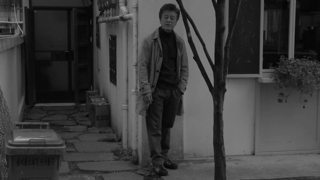 Walk Up (2022) movie still: fullbody — An outdoor space next to a white building with exposed piping and trash bins. A middle-aged man stands…
