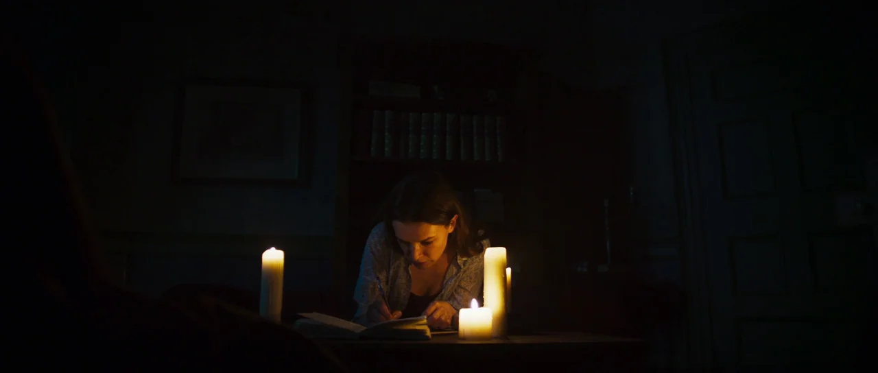 A Dark Song (2016) movie still: medium — A dimly lit room features a bookshelf filled with volumes in the background. A woman sits at a wooden table…