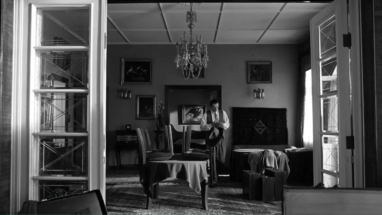 Schindler's List (1993) movie still: medium — A furnished hotel room features a central dining table with chairs and a chandelier. A man in a vest…