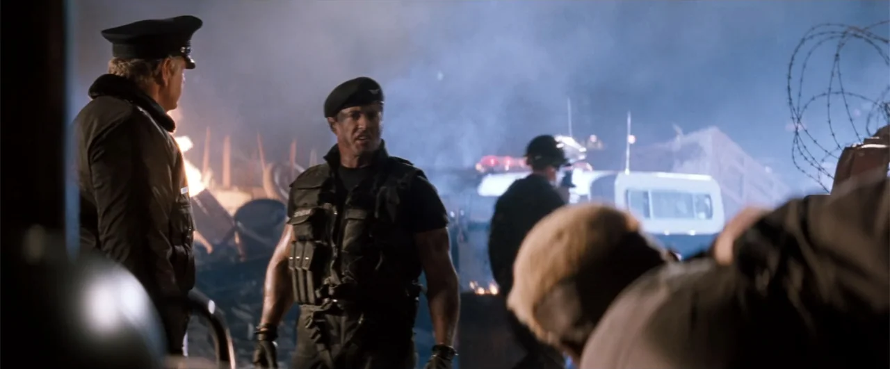 Demolition Man (1993) movie still: medium — An outdoor industrial site at night features piles of rubble and smoke-filled air. A man in a tactical…