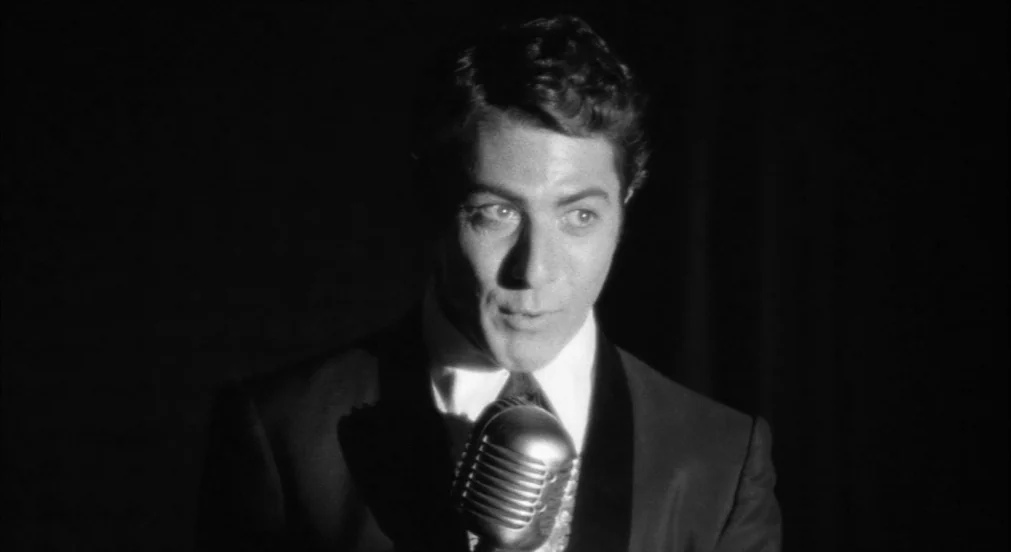 Lenny (1974) movie still: closeup — A dark stage with an empty background behind a performer. A man stands at a microphone stand illuminated by a…