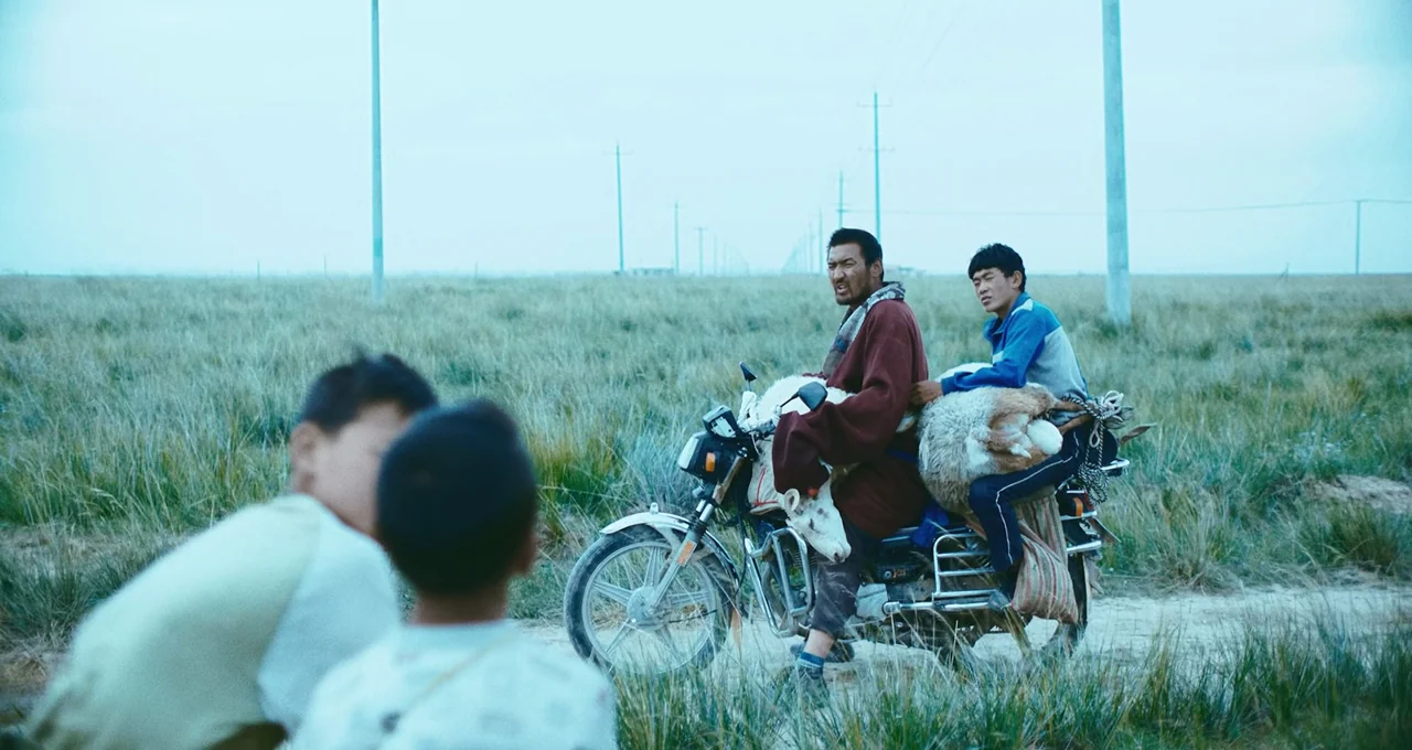 Balloon (2019) movie still: wide — Two men ride a motorcycle through a vast, flat grassland under an overcast sky. They are carrying a sheep across…
