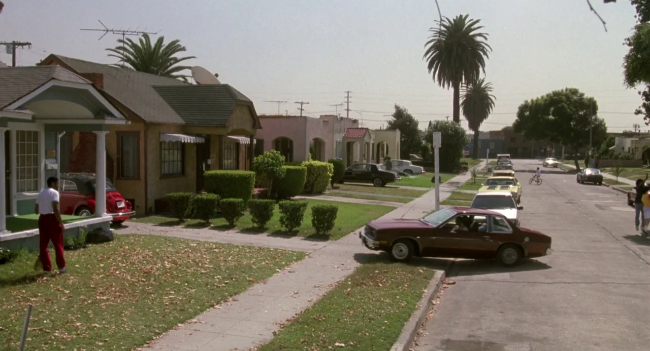 Boyz In The Hood (1991) movie still: wide — A residential neighborhood street with stucco houses and palm trees during a sunny day. A man in red…