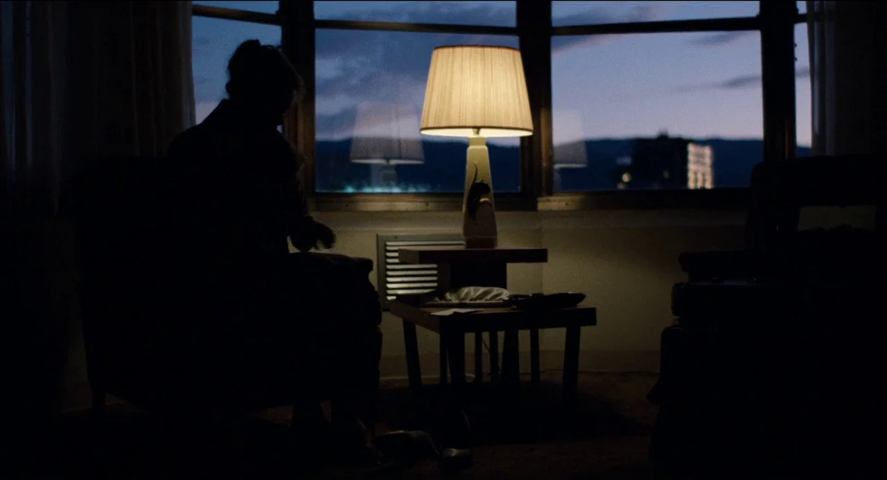 Desert Hearts (1985) movie still: medium — A dark hotel room features a large window overlooking a dusk cityscape. A woman sits in a chair,…