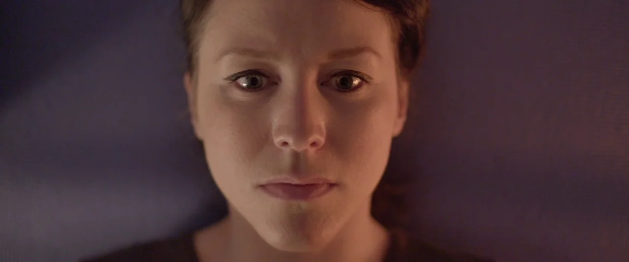 Prevenge (2016) movie still: closeup — A close-up shot focuses on a woman's face against a dark purple backdrop. She looks directly into the camera…