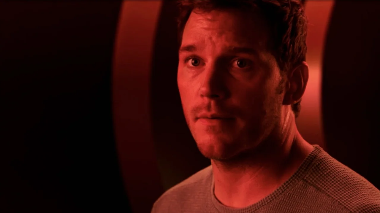 Passengers (2016) movie still: closeup — A darkened interior space with minimal architectural detail and a red light source. A man in a grey shirt…
